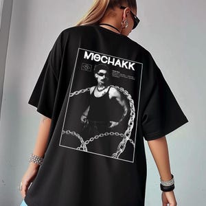 Puede incluir: Camiseta negra de gran tamaño con estampado gráfico en la espalda. El estampado incluye la palabra "MOCHAKK" sobre una imagen en blanco y negro de un hombre con camiseta sin mangas y gafas de sol, rodeado de cadenas.