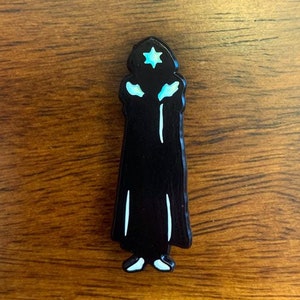 Puede incluir: Pin de esmalte negro de una persona que lleva una t&uacute;nica negra y una estrella azul en la cabeza.
