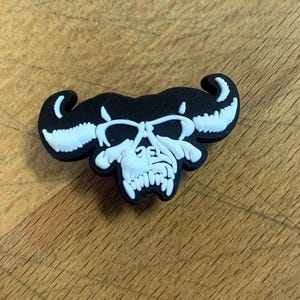 Może przedstawiać: Czarno-biały urok czaszki z rogami, przypominający głowę byka. Urok jest wykonany z gumy i ma błyszczące wykończenie.