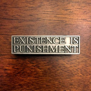 Può includere: Uno spillo in metallo argentato con il testo "EXISTENCE IS PUNISHMENT" inciso su di esso.