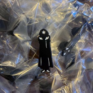 Puede incluir: Un pin de esmalte negro con la silueta de una persona que lleva una t&uacute;nica y una estrella en la cabeza. El pin est&aacute; en una bolsa de pl&aacute;stico transparente.