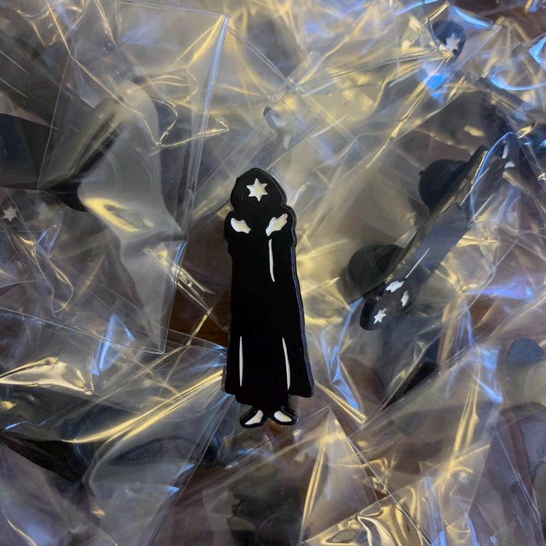 Puede incluir: Un pin de esmalte negro con la silueta de una persona que lleva una t&uacute;nica y una estrella en la cabeza. El pin est&aacute; en una bolsa de pl&aacute;stico transparente.
