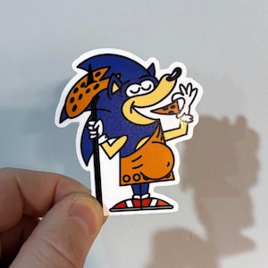 May include: A sticker featuring a cartoon character resembling Sonic the Hedgehog, depicted as a caveman. The character is blue with a yellow snout and orange clothing, holding a pizza slice and a pizza-topped spear. The sticker has a white border.