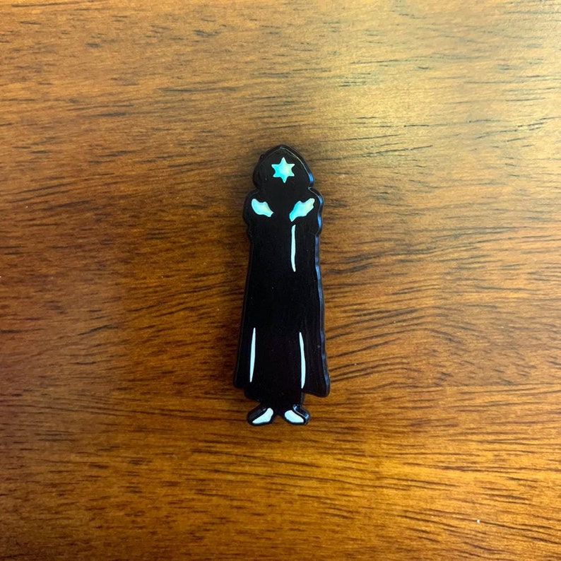 Puede incluir: Pin de esmalte negro de una persona que lleva una t&uacute;nica negra y una estrella azul en la cabeza.