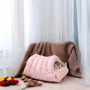 May include: A pink and white patterned cat bed with a cat sleeping inside. The bed is shaped like a tunnel and has a soft, plush lining.