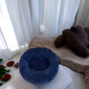 以下が含まれることがあります： 丸い形をした、柔らかくふわふわの青いぬいぐるみ製のペットベッド。ベッドは白い表面の上に置かれています。