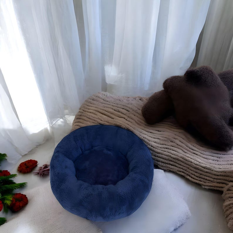以下が含まれることがあります： 丸い形をした、柔らかくふわふわの青いぬいぐるみ製のペットベッド。ベッドは白い表面の上に置かれています。