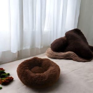 以下が含まれることがあります： 茶色のふわふわしたペットベッドが2つ。1つは縁が高くなった丸いベッド、もう1つは折り畳まれた縁のある長方形のベッドです。