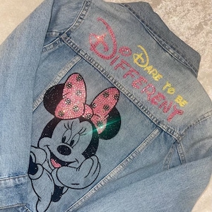 May include: Light blue denim jacket featuring a Minnie Mouse graphic with a pink bow and the text "Dare to be Different". The Disney-themed jacket has rhinestone embellishments.
