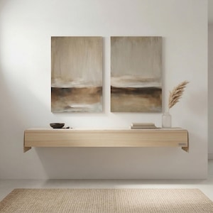 May include: Two abstract landscape paintings in neutral tones hang above a light wood floating shelf. The paintings feature a blend of beige, brown, and grey hues. A vase with dried plants and a small bowl are on the shelf.