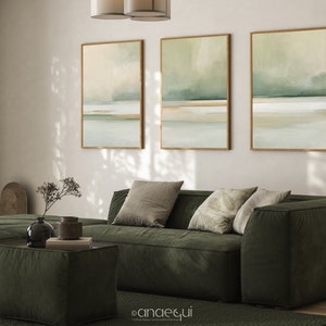 May include: Three framed abstract landscape art prints in shades of green, beige, and white hang above a dark green sofa with throw pillows. A matching ottoman and rug complete the living room decor. The artwork features a minimalist design.