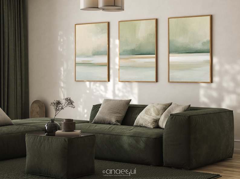 May include: Three framed abstract landscape art prints in shades of green, beige, and white hang above a dark green sofa with throw pillows. A matching ottoman and rug complete the living room decor. The artwork features a minimalist design.