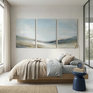 May include: A bedroom scene featuring a bed with beige bedding, a light blue duvet, and several pillows. Above the bed are three abstract landscape paintings in shades of blue, beige, and brown. A blue side table holds a clock and books.