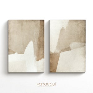 May include: Two abstract art prints. Each print features a beige and white color palette with brushstroke-style shapes. The artwork is displayed on a white background. The prints are rectangular and appear to be canvas.