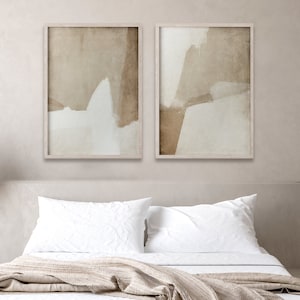 May include: Two framed abstract art prints. Each print features a neutral color palette with beige, brown, and white brushstrokes. The artwork is displayed above a bed with white linens and a beige blanket, creating a calming bedroom aesthetic.