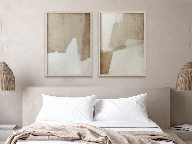 May include: Two framed abstract art prints. Each print features a neutral color palette with beige, brown, and white brushstrokes. The artwork is displayed above a bed with white linens and a beige blanket, creating a calming bedroom aesthetic.