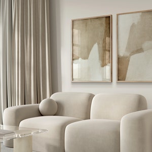 May include: A modern living room featuring a cream-colored sofa, two abstract art prints in wooden frames, and a minimalist floor lamp. The artwork displays neutral tones of beige and white. Soft, neutral-toned curtains frame the scene.