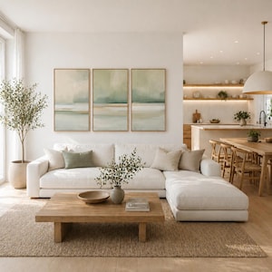 May include: A modern living room with a white sectional sofa, a wooden coffee table, and three framed landscape paintings. A large area rug and a potted tree add warmth. The kitchen and dining area are visible in the background.