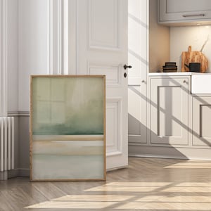 May include: A framed abstract art print with a light wood frame. The artwork features a watercolor-style landscape in shades of green, beige, and blue. The print is leaning against a white door in a room with hardwood floors.