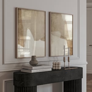 May include: Two framed abstract art prints with neutral tones hang above a black console table. The table displays a vase, books, a cube candle, and two brass candle holders with white candles. The room has a minimalist aesthetic.