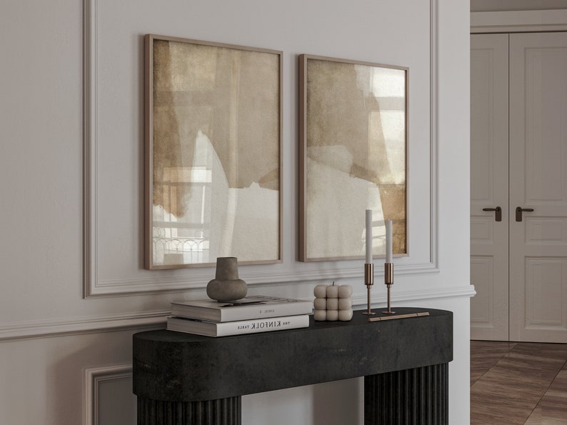May include: Two framed abstract art prints with neutral tones hang above a black console table. The table displays a vase, books, a cube candle, and two brass candle holders with white candles. The room has a minimalist aesthetic.