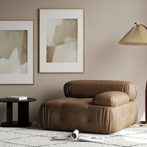May include: A modern living room scene featuring a brown leather armchair, a black side table, and a floor lamp with a pleated lampshade. Two framed abstract art pieces hang on the wall. A patterned rug and open magazine complete the decor.