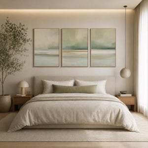 May include: A bedroom scene featuring a bed with neutral-toned bedding, a large headboard, and a long green bolster pillow. Above the bed are three framed abstract landscape paintings in muted colors. A small side table with a lamp and a potted plant are also visible.