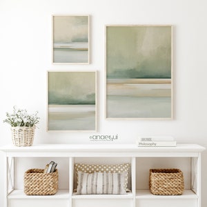 May include: Three framed abstract landscape art prints in muted green, beige, and gray tones. The artwork is displayed above a white shelf with woven baskets, a decorative pillow, and a small floral arrangement in a basket.
