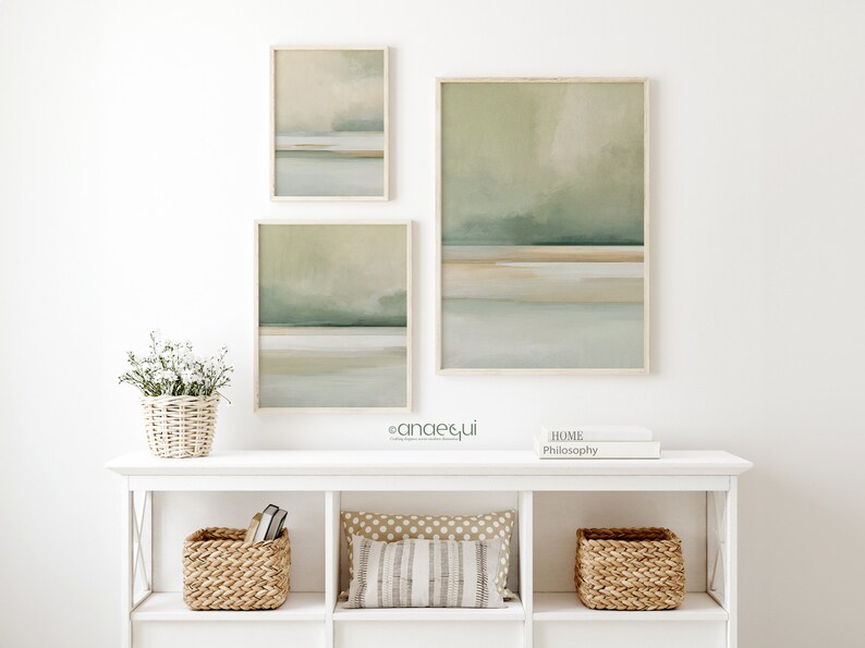 May include: Three framed abstract landscape art prints in muted green, beige, and gray tones. The artwork is displayed above a white shelf with woven baskets, a decorative pillow, and a small floral arrangement in a basket.
