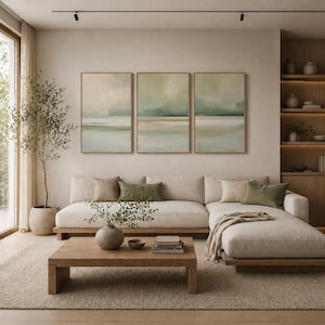 May include: A modern living room with a neutral color palette. A large beige sectional sofa with decorative pillows faces a wooden coffee table. Three framed abstract landscape paintings hang above the sofa. A potted tree and built-in shelving complete the space.