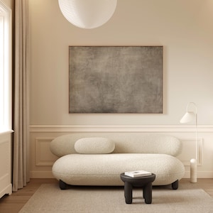 May include: A modern living room featuring a cream-colored boucle sofa with a matching bolster pillow. A small, dark gray side table holds a book. A large abstract painting hangs above the sofa, and a white floor lamp stands nearby.