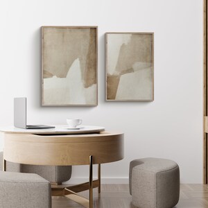 May include: Two framed abstract art prints with neutral tones of beige and white hang on a white wall. The artwork is displayed above a round wooden table with a laptop and a teacup. Two upholstered ottomans are in front of the table.