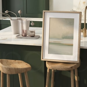 May include: A framed abstract landscape art print with a light wood frame rests on a wooden stool. The artwork features soft, muted colors of beige, green, and blue. The kitchen setting includes a white countertop, green cabinets, and a gold faucet.
