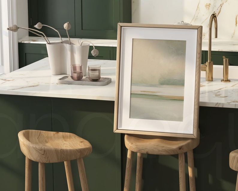 May include: A framed abstract landscape art print with a light wood frame rests on a wooden stool. The artwork features soft, muted colors of beige, green, and blue. The kitchen setting includes a white countertop, green cabinets, and a gold faucet.