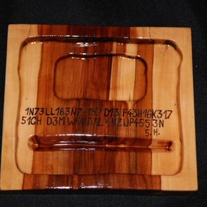 May include: A wooden tray with a rectangular cutout in the center. The tray is made of light and dark wood and has a glossy finish. The text "1N73 LL163NZ 157 D13 F43H16K317 51GH D3M WAND3L 4NZ UP4-553N 5.H." is engraved on the tray.