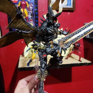 Pode incluir: Figura de ação detalhada de um monstro preto e dourado com grandes asas e uma espada. A figura tem um design pontiagudo e está em pose com sua espada levantada. Outras figuras de ação são visíveis no fundo.
