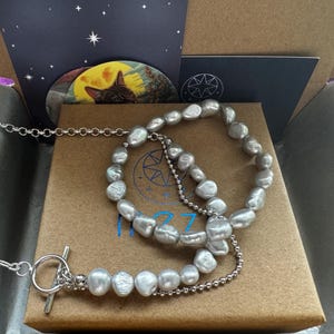 May include: A silver chain necklace with a toggle clasp and a silver chain bracelet with a silver chain extender. Both are adorned with a strand of large, irregular, gray pearls.