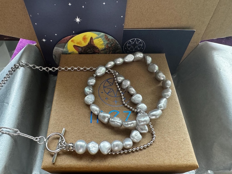 May include: A silver chain necklace with a toggle clasp and a silver chain bracelet with a silver chain extender. Both are adorned with a strand of large, irregular, gray pearls.