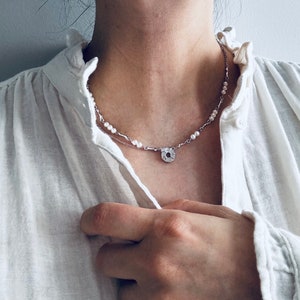 Può includere: Una collana d'argento con una perla e un ciondolo circolare. La collana è indossata da una persona che indossa una camicia bianca.