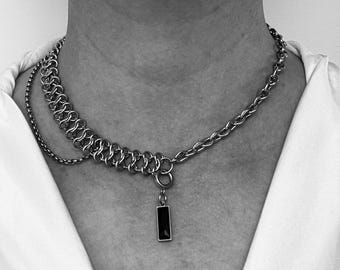 Puede incluir: Un collar de cadena de plata con un colgante rectangular negro. El collar está formado por dos cadenas de estilos diferentes.