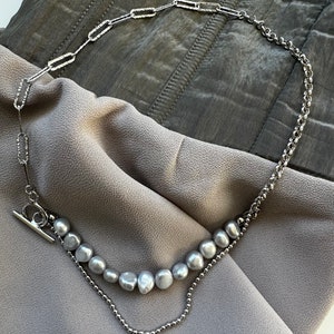 May include: A silver chain necklace with a silver toggle clasp and a strand of gray pearls.