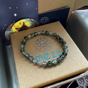May include: A green and gray beaded bracelet with a silver clasp. The bracelet is in a brown box with the word "mezs" printed on it.