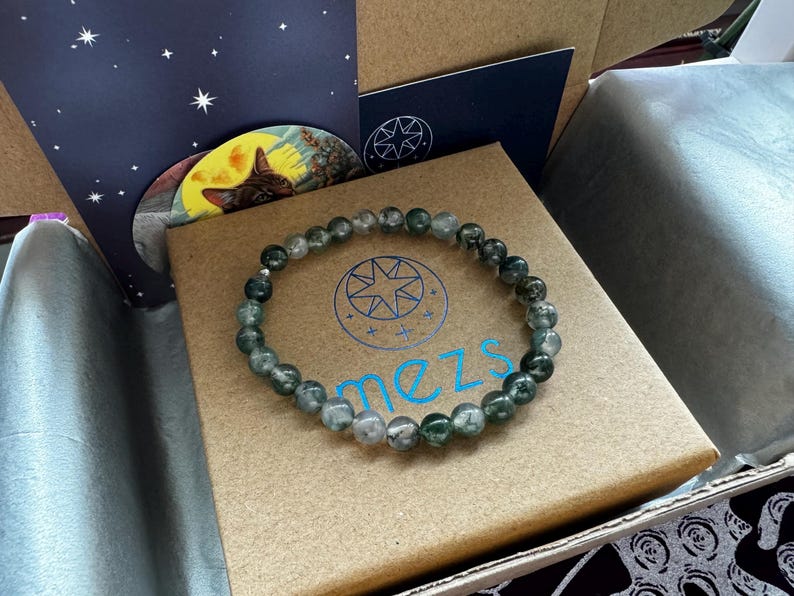 May include: A green and gray beaded bracelet with a silver clasp. The bracelet is in a brown box with the word "mezs" printed on it.