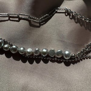 May include: A silver chain necklace with a row of large, gray pearls. The necklace is layered with a smaller silver chain.