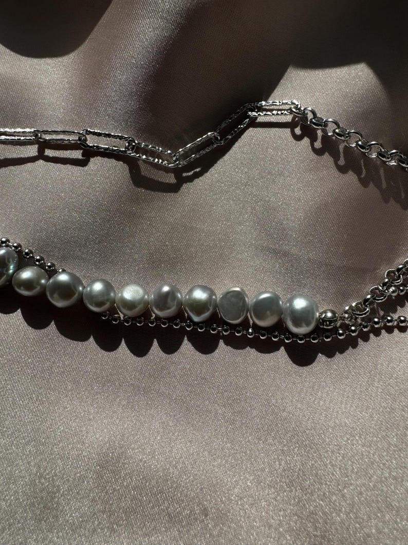 May include: A silver chain necklace with a row of large, gray pearls. The necklace is layered with a smaller silver chain.
