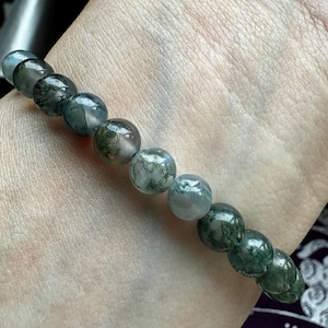 May include: A green moss agate bracelet with round beads. The bracelet is on a person's wrist.