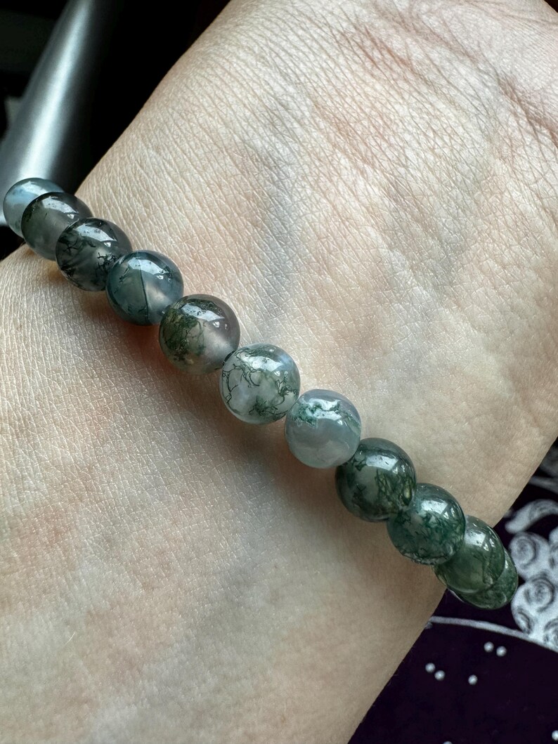 May include: A green moss agate bracelet with round beads. The bracelet is on a person's wrist.