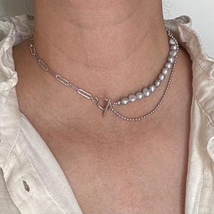 May include: A silver chain necklace with a silver chain and a strand of faux pearls. The necklace is layered and has a toggle clasp.