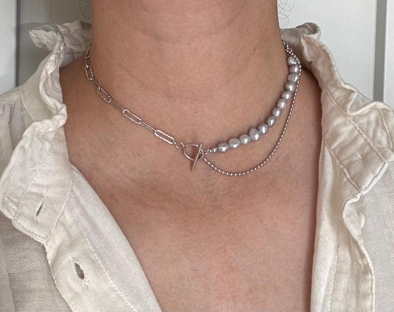 May include: A silver chain necklace with a silver chain and a strand of faux pearls. The necklace is layered and has a toggle clasp.
