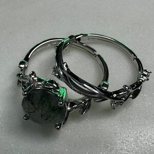 May include: A silver ring set with a large, dark green gemstone. The ring features a delicate, leafy design.
